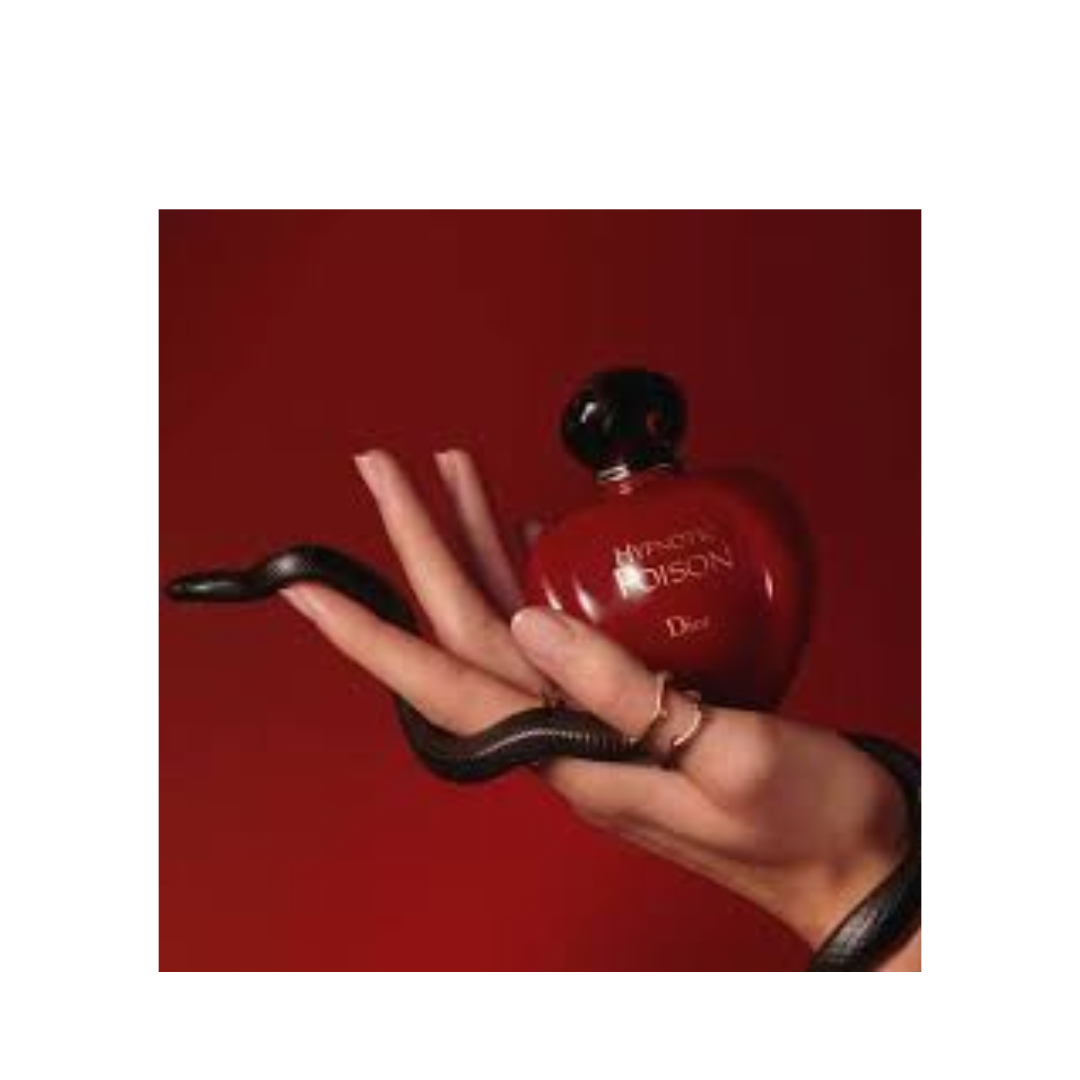 Dior Miracle Poison perfume bottle in a glossy red heart shape with a black cap, held in a hand with a black snake draped over the hand, against a solid red background.