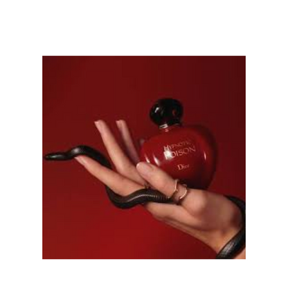 Dior Miracle Poison perfume bottle in a glossy red heart shape with a black cap, held in a hand with a black snake draped over the hand, against a solid red background.