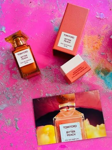 Tom Ford Bitter Peach Eau de Parfum set on a vibrant pink background, featuring the amber glass bottle, its coral pink box, a small white and coral pink sample vial, and a printed card depicting the bottle.