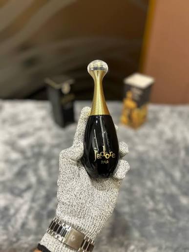 A hand wearing a gray knit glove holds a Dior Addict perfume bottle. The bottle is glossy black with a metallic gold neck and a clear spherical cap.