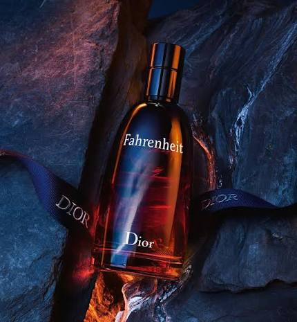 Dior Fahrenheit perfume bottle with amber liquid, displayed on dark rocks with a branded ribbon. The bottle has a black cap and features 'Fahrenheit' and 'Dior' branding.