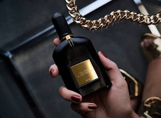 Hand holding a black and gold perfume bottle with a gold chain in the background