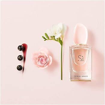 A bottle of Giorgio Armani Si perfume with a pale pink cap and clear glass, styled on a light pink background with a pink rose, a white flower, and three dark berries.